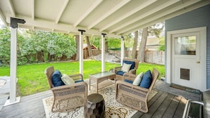Un patio techado con muebles de mimbre y una pequeña mesa, una terraza de madera y vistas a un patio trasero con valla de madera y árboles.