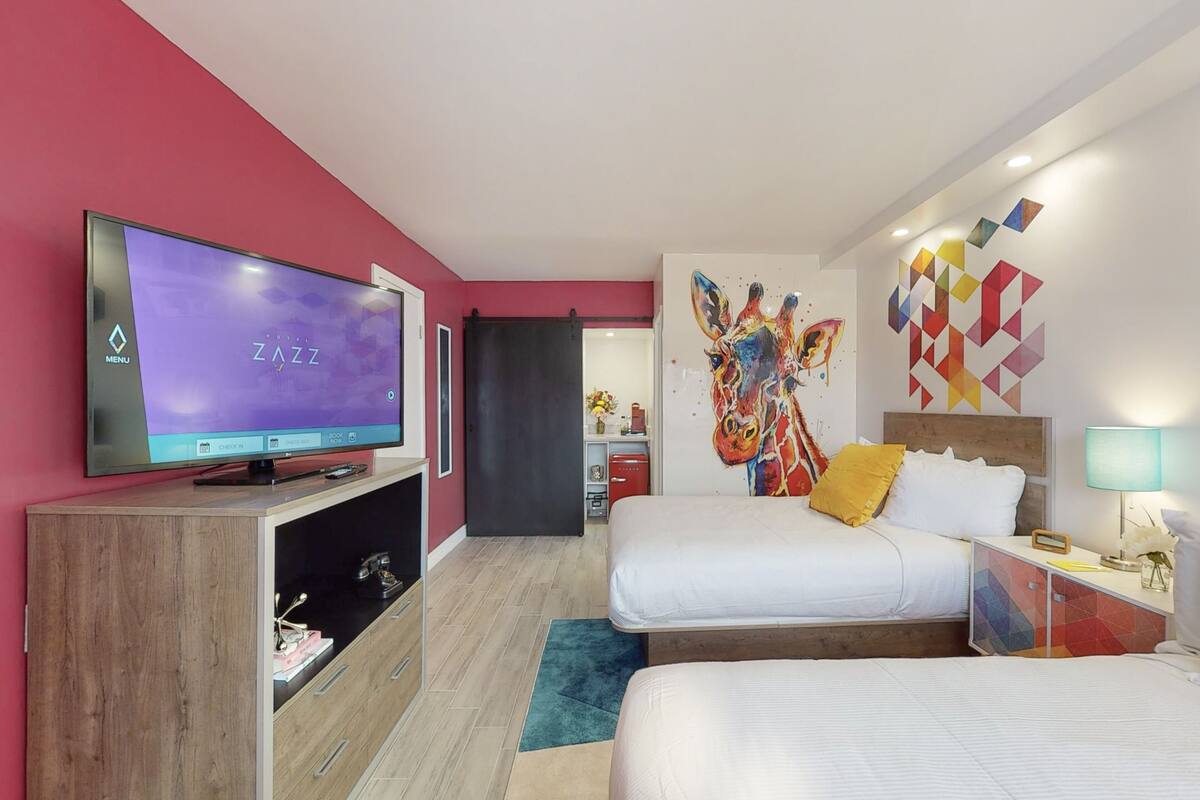 A modern hotel room with a large flat-screen TV, two beds, a colorful abstract wall art, and a wooden cabinet.