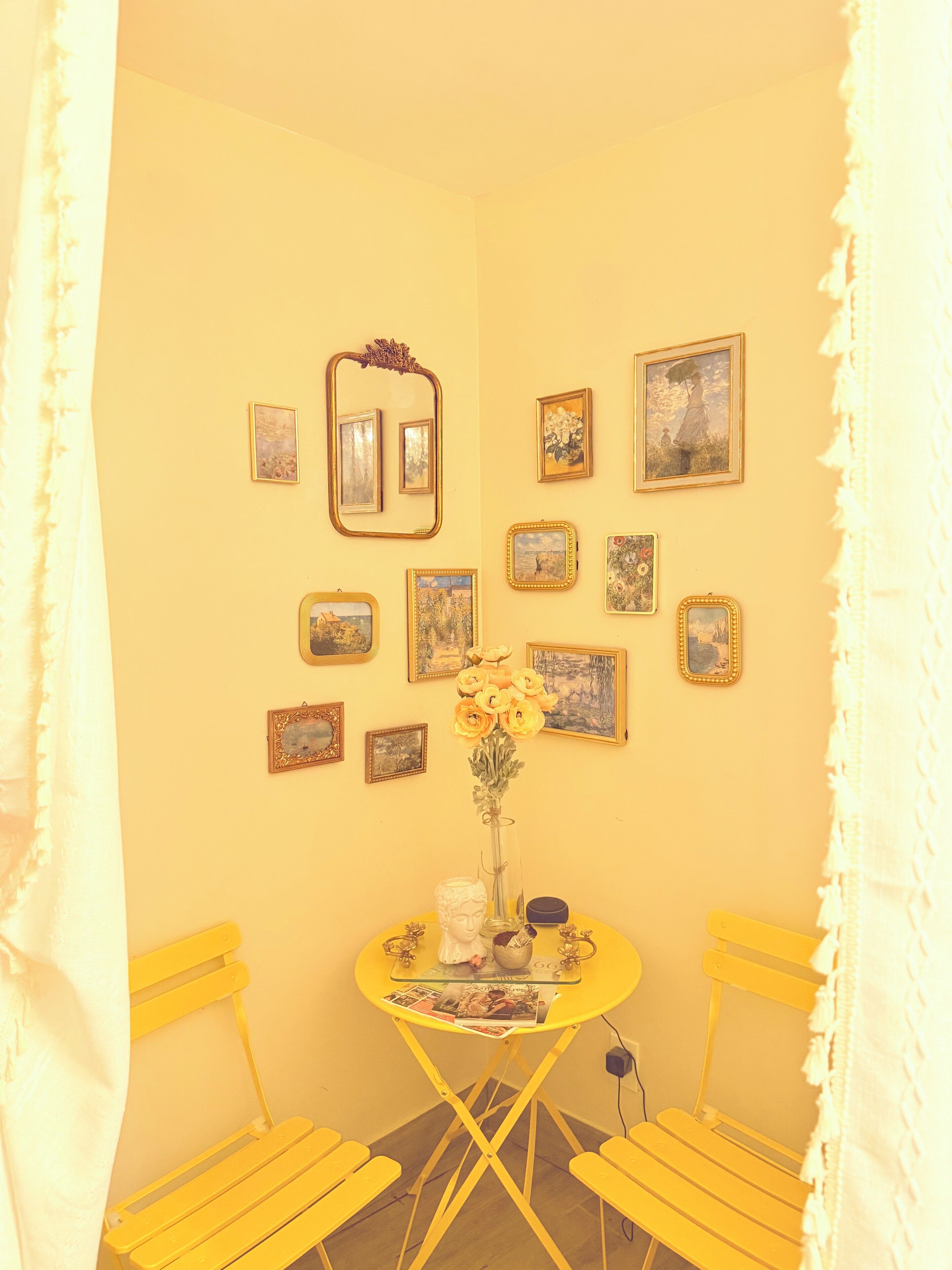 A room with a yellow wall, a round table with a vase of flowers, and several framed pictures. There are two yellow chairs and a white curtain.