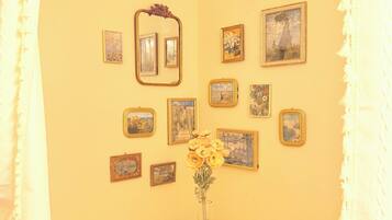 A room with a yellow wall, a round table with a vase of flowers, and several framed pictures. There are two yellow chairs and a white curtain.