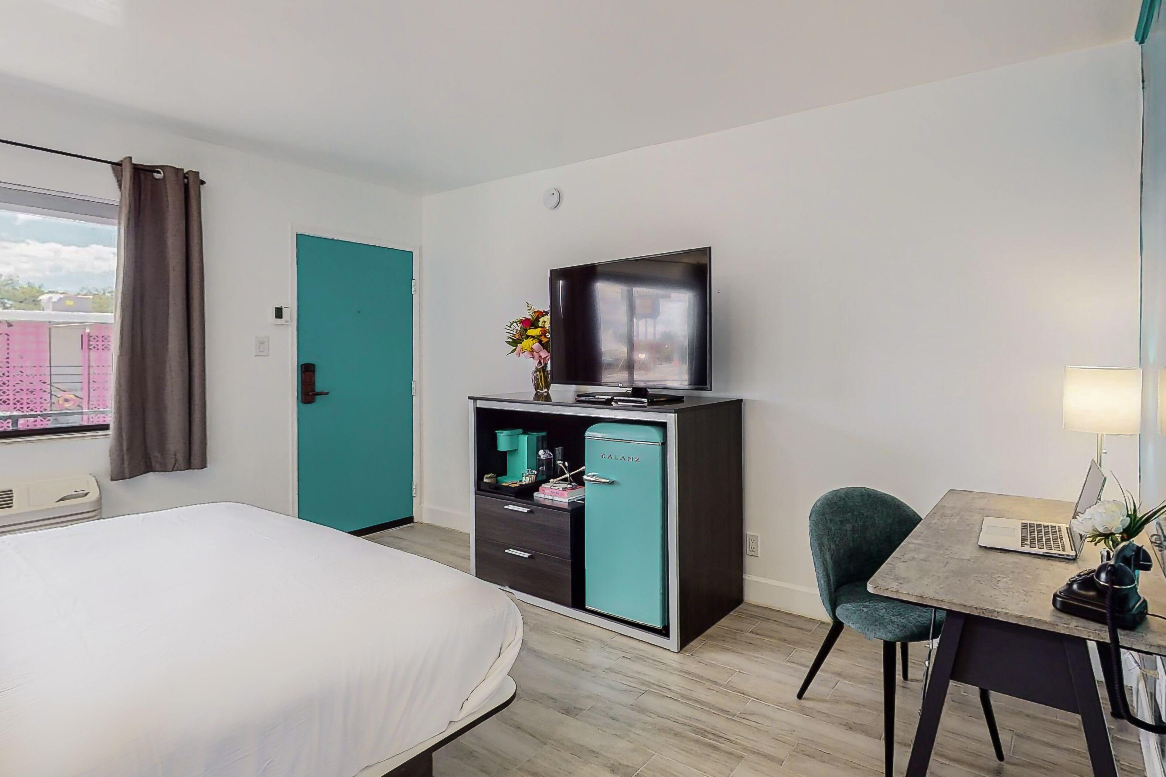 A hotel room with a bed, a desk with a laptop, a television, a teal-colored minibar, and a window with curtains.