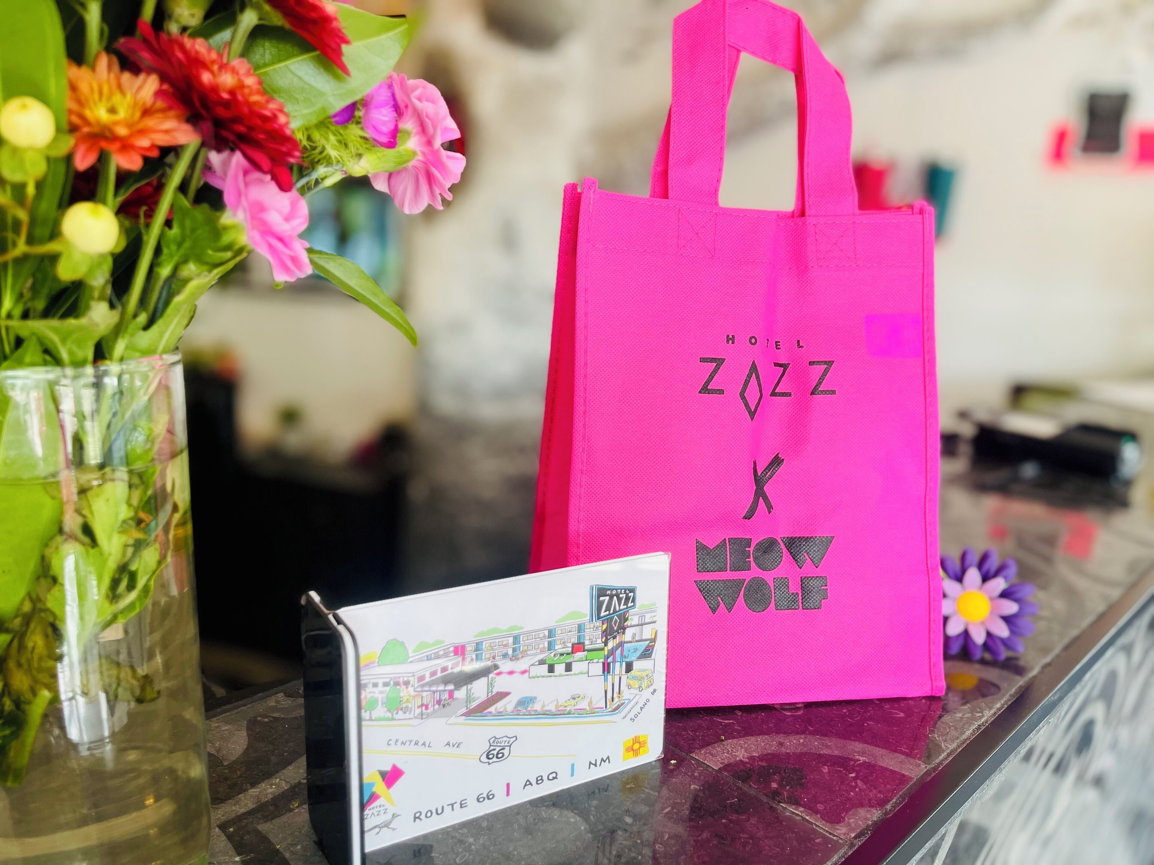 Una borsa rosa da albergo con la scritta "Hotel Zazz" e "Knew Wolf", un vaso di vetro con fiori e una mappa stampata della Route 66.