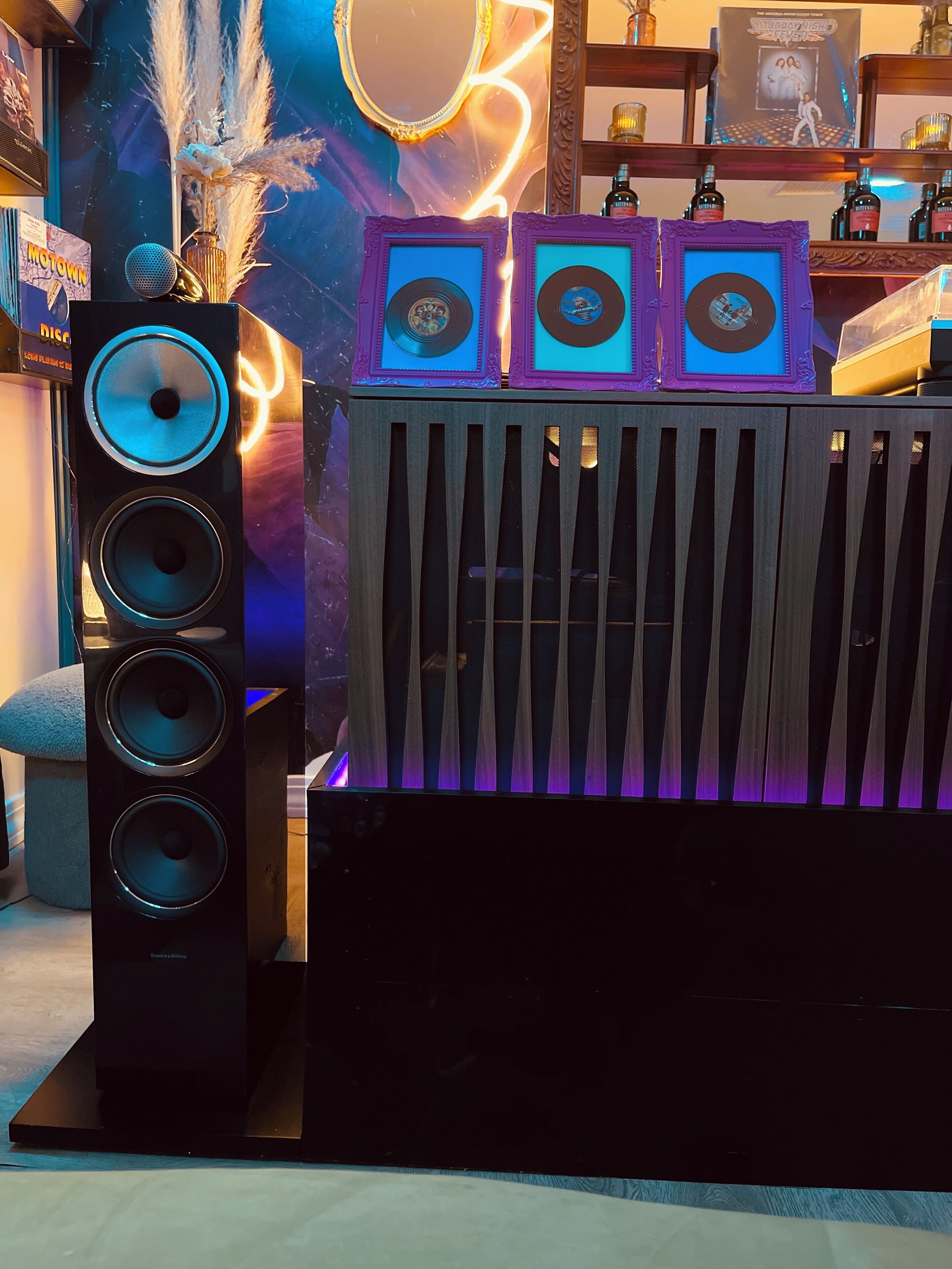 Sistema de som com vários alto-falantes e um subwoofer, um letreiro de neon e discos de vinil emoldurados numa estante.