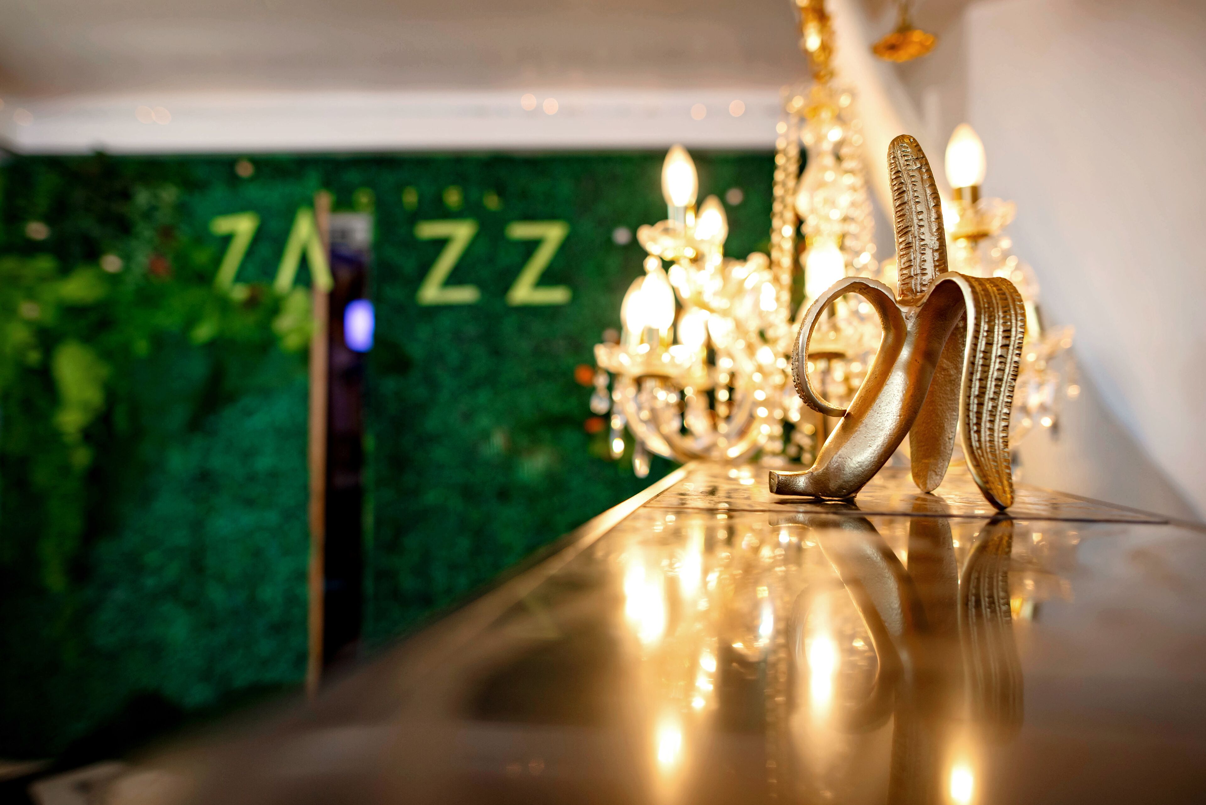 A golden shoe on a reflective surface with a chandelier and a green wall with 'ZZZ' in the background.