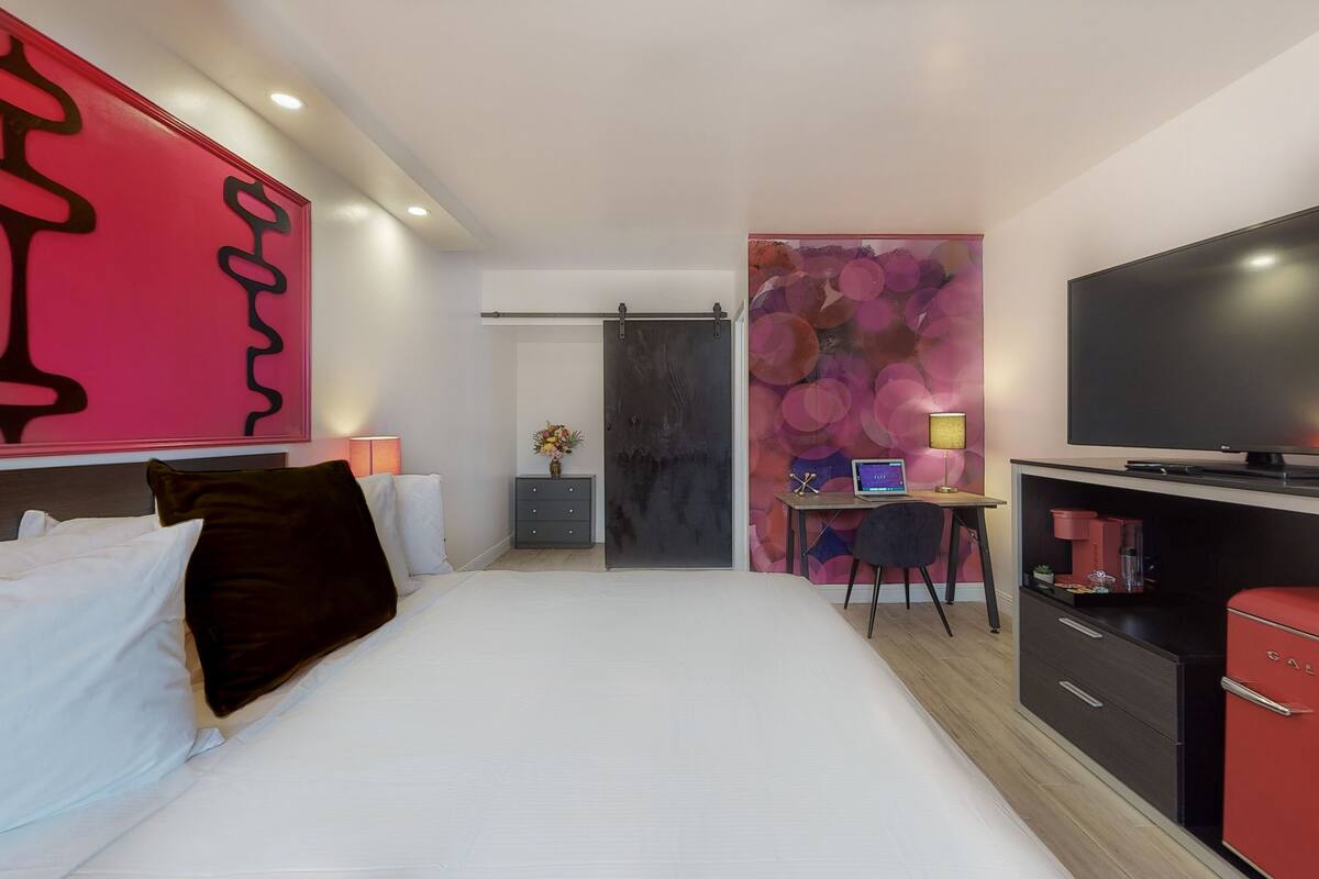 A modern hotel room with a bed, a desk with a computer, a TV, and a red appliance.