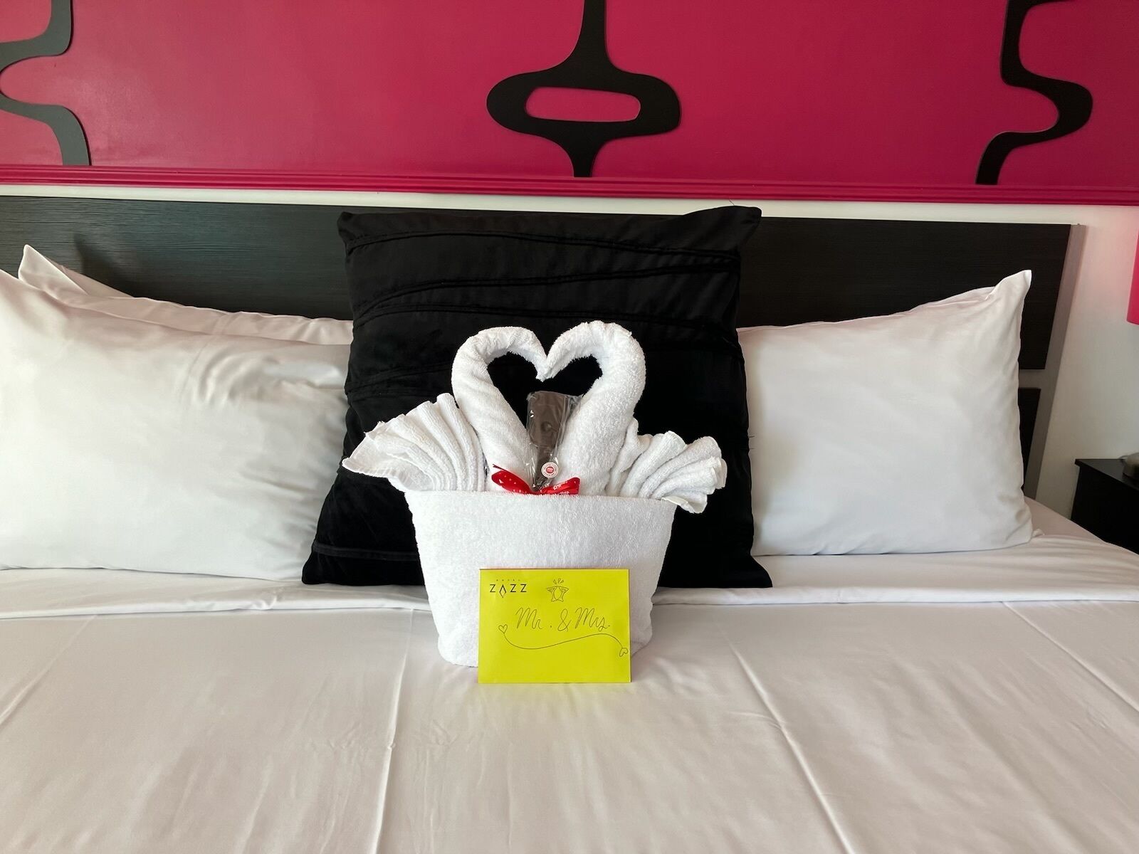 A hotel room with a bed, white pillows, a black pillow, a heart-shaped towel arrangement, and a yellow sign with text.