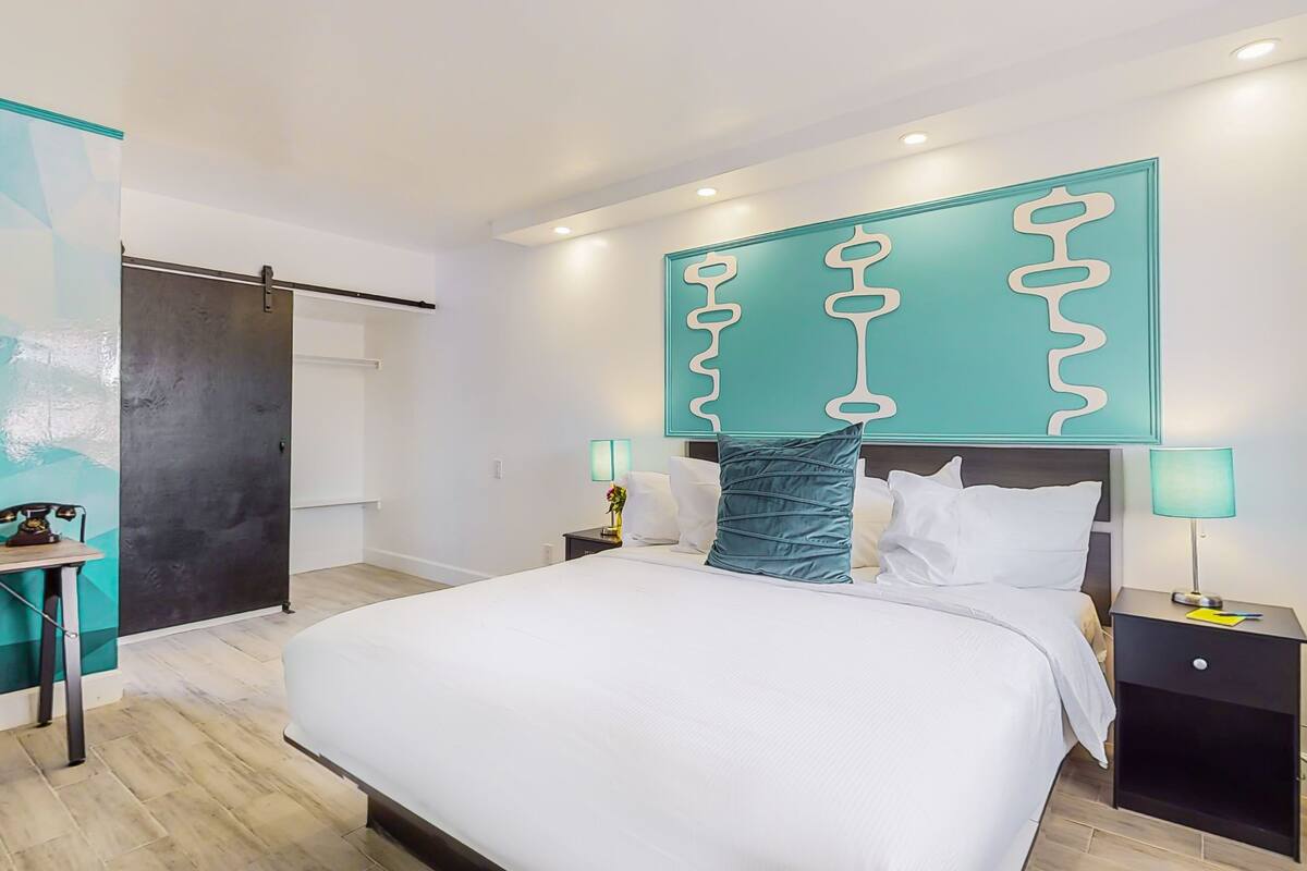 A modern hotel room with a large bed, a desk, and a wall-mounted artwork.