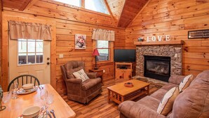 Un intérieur de chalet chaleureux avec des murs en bois, une cheminée en pierre, un téléviseur, une table à manger et un coin salon confortable.