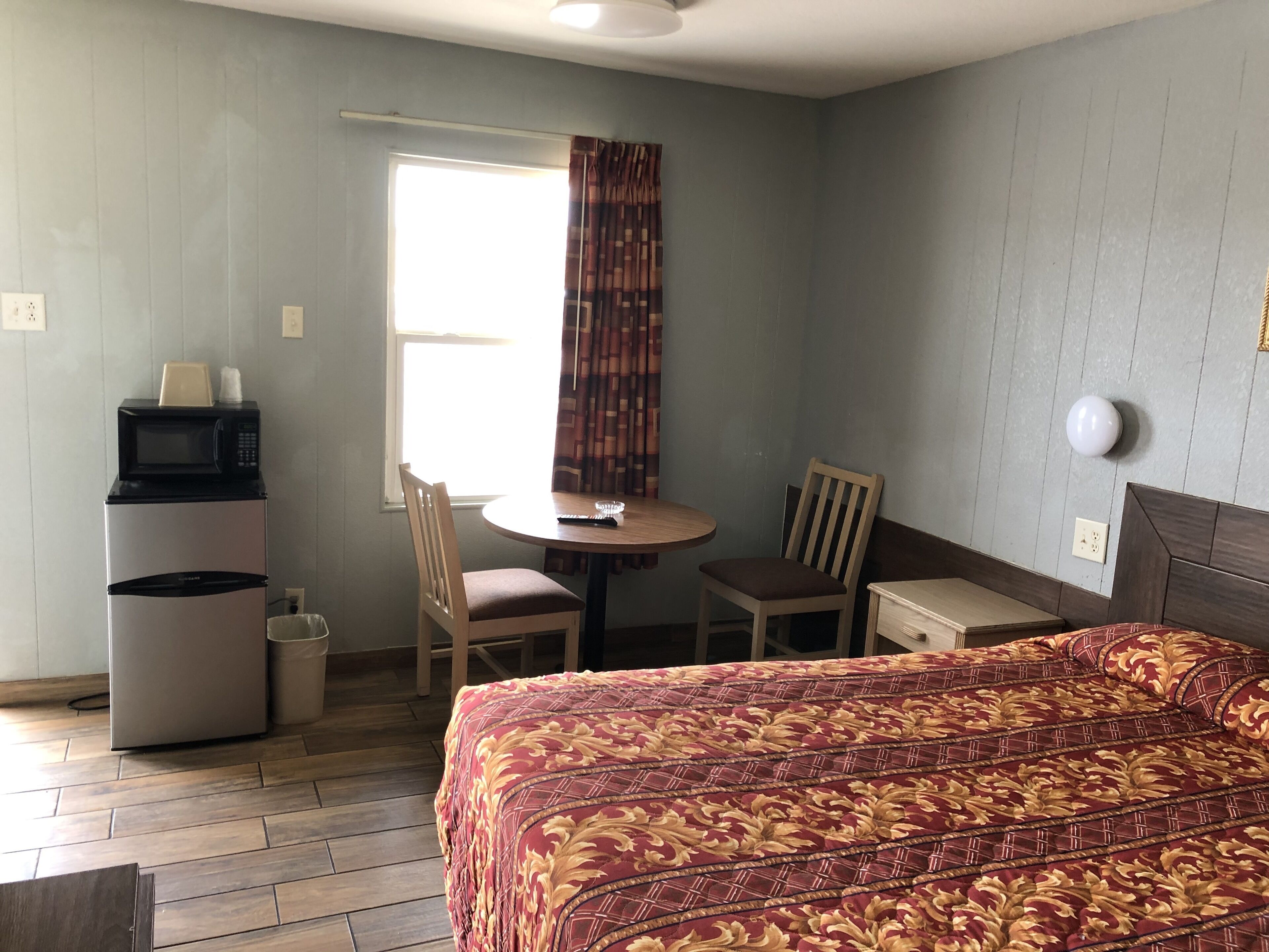 A hotel room with a bed, a desk with a chair, a microwave, and a window with curtains.