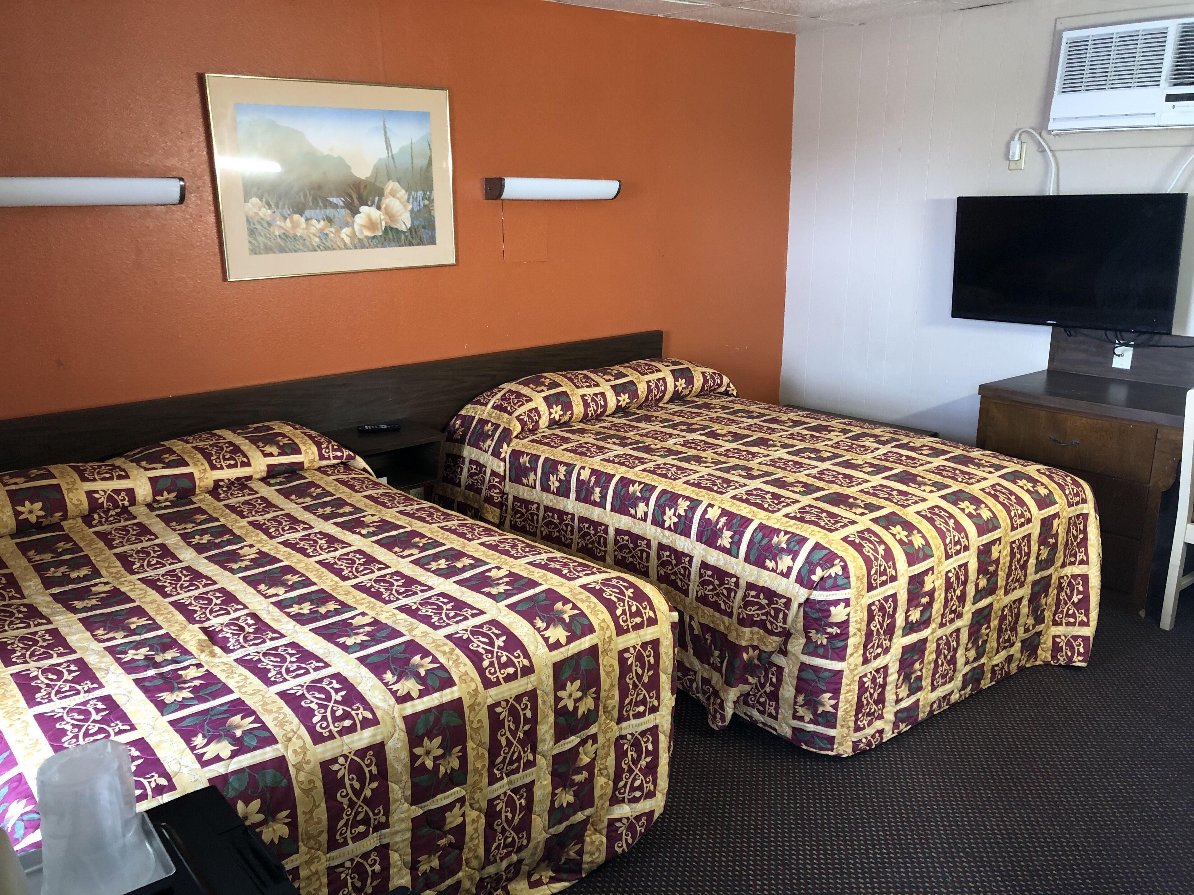 A hotel room with two beds, a flat-screen TV, a painting on the wall, and an air conditioning unit.