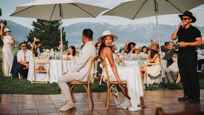 An outdoor wedding reception with guests seated at tables under umbrellas, with a mountainous backdrop.
