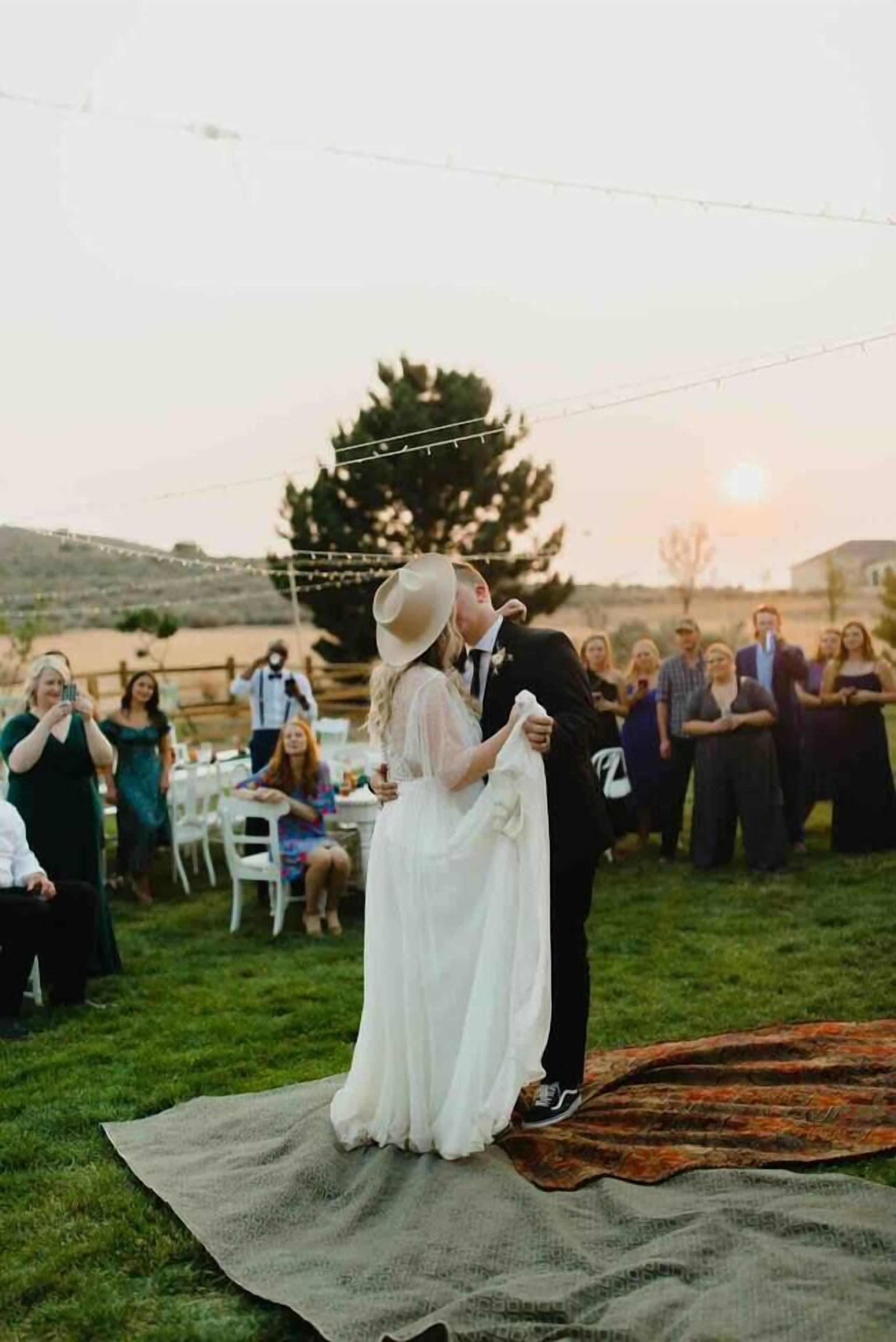 A wedding ceremony with a couple embracing on a blanket, guests seated around them, and a sunset in the background.