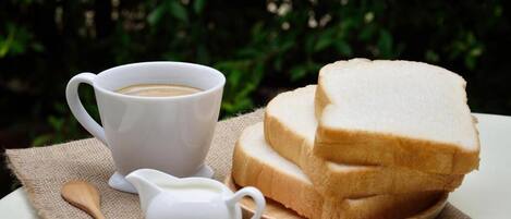 Una taza de café blanco, una pequeña jarra blanca y una pila de pan blanco sobre un plato de madera, todo sobre una mesa con mantel de arpillera.