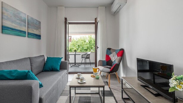 A modern living room with a grey sofa, a colorful armchair, a coffee table, and a flat-screen TV. There is a balcony with a table and chairs, and a view of greenery outside. - Oroz / Apartment Oroz I (Porec)