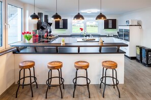 A modern kitchen with a central island, bar stools, pendant lighting, and a view of the outdoors. - Hotel Sauerlandvilla Winterberg (Winterberg)