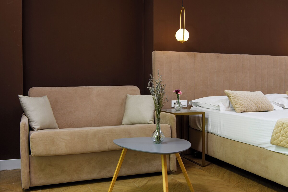 Una habitación con sofá, una mesita redonda con un florero, una mesita de noche con lámpara y una cama con almohadas.
