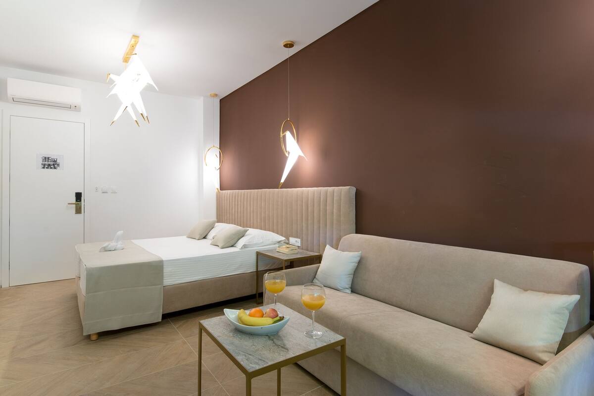 A modern hotel room with a bed, a sofa, a small table with fruit and drinks, and unique lighting fixtures.
