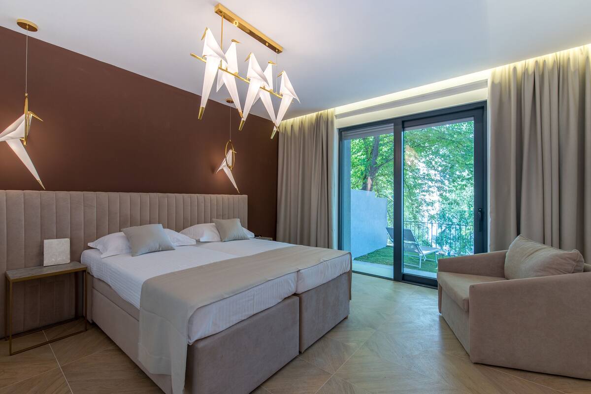 A modern bedroom with a large bed, a nightstand, a sofa, and a view of the outdoors through a sliding glass door.