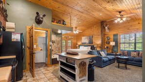 L’intérieur d’un chalet rustique présente un plafond en bois, un coin repas avec un îlot central, un salon avec un canapé bleu et une cuisine dotée d’un réfrigérateur noir.
