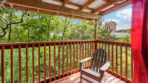 Une terrasse en bois avec une rambarde rouge, une seule chaise Adirondack et une vue sur des arbres verdoyants et un bâtiment situé au loin.