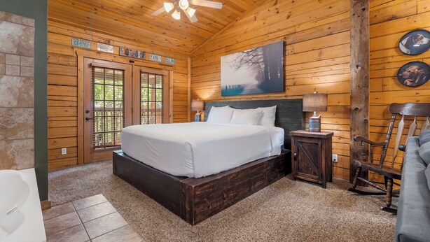 Une chambre à coucher située dans une cabane en bois, avec un lit, un ventilateur de plafond, une table de chevet, un fauteuil à bascule et un tableau accroché au mur.