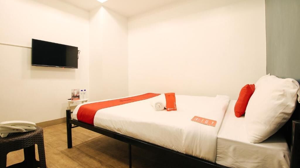 Un lit simple avec une literie blanche et rouge, un téléviseur à écran plat fixé au mur, une table de chevet avec un téléphone et un petit tabouret.