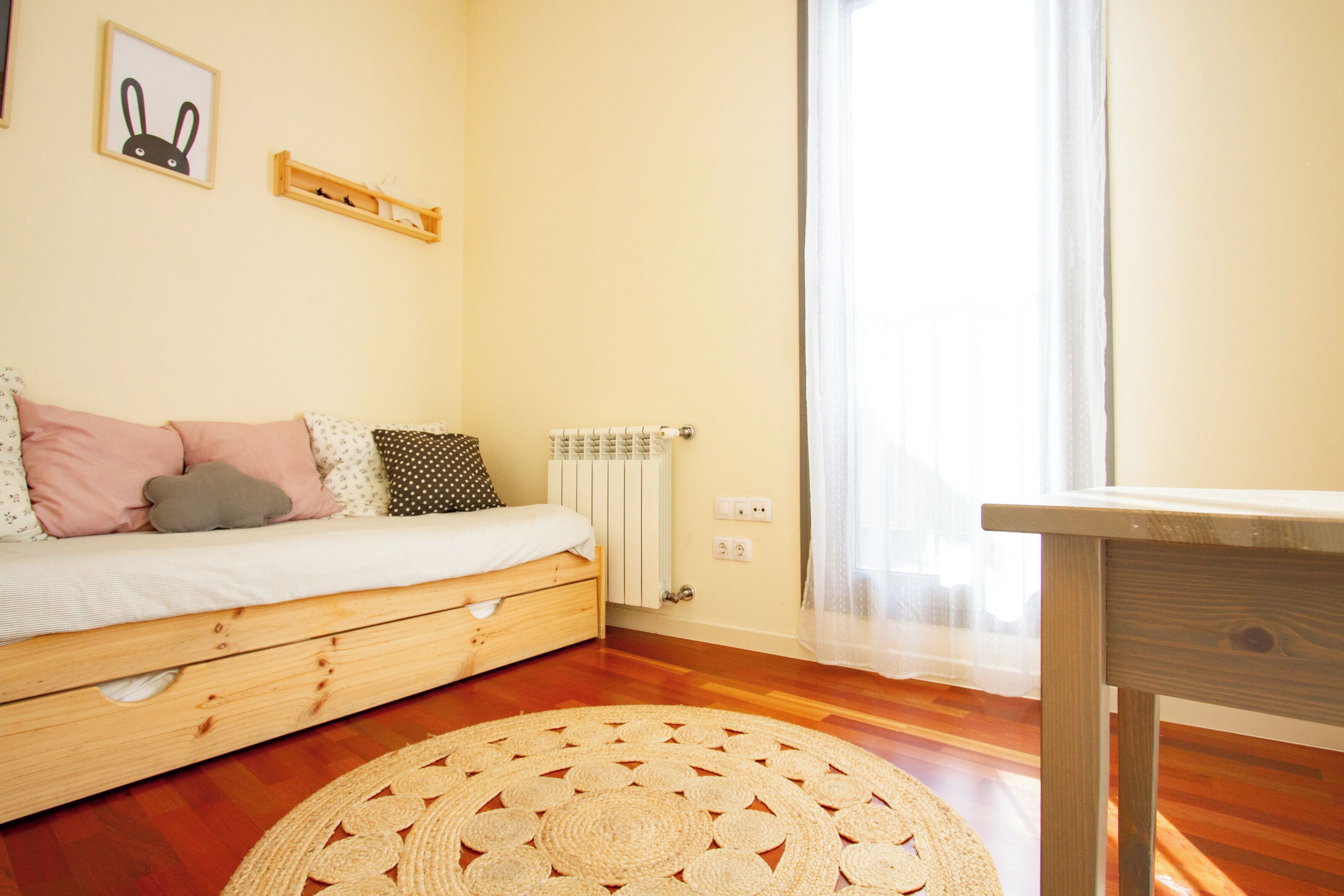 A bedroom with a wooden bed, a round woven rug, a radiator, and a window with sheer curtains.