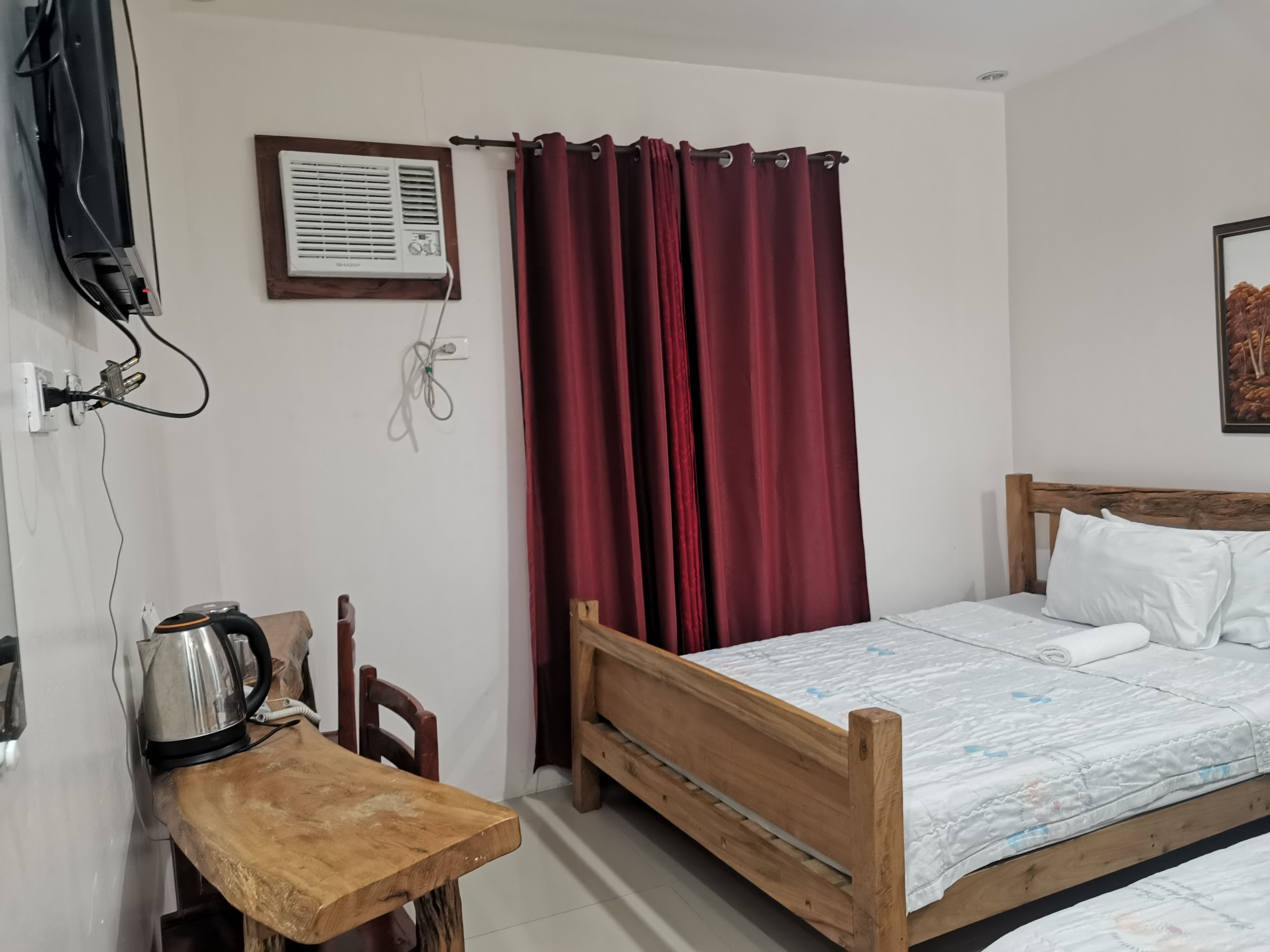 Ein Schlafzimmer mit einem hölzernen Bett, weißen Kissen, einem Nachttisch aus Holz mit einem Wasserkocher, einem roten Vorhang und einer Wand-Klimaanlage.
