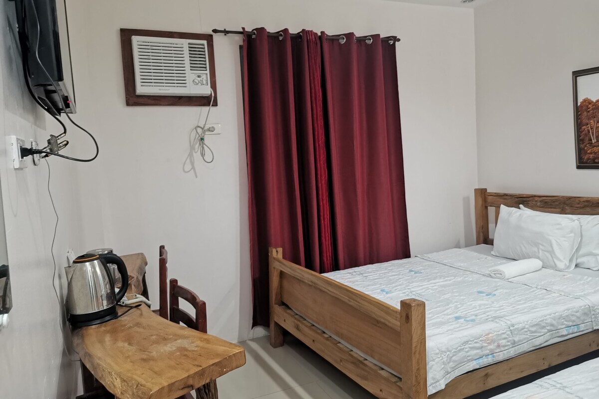 Un dormitorio con una cama de madera, almohadas blancas, una mesita de noche de madera con una tetera, una cortina roja y un aire acondicionado instalado en la pared.