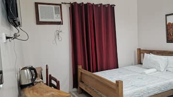 Une chambre avec un lit en bois, des oreillers blancs, une table de chevet en bois avec une bouilloire, un rideau rouge et un climatiseur fixé au mur.