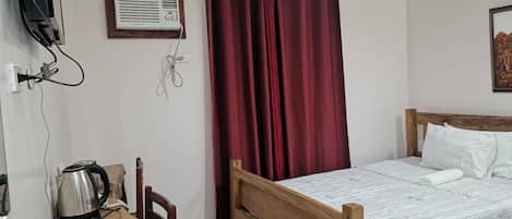 A bedroom with a wooden bed, white pillows, a wooden nightstand with a kettle, a red curtain, and a wall-mounted air conditioner.