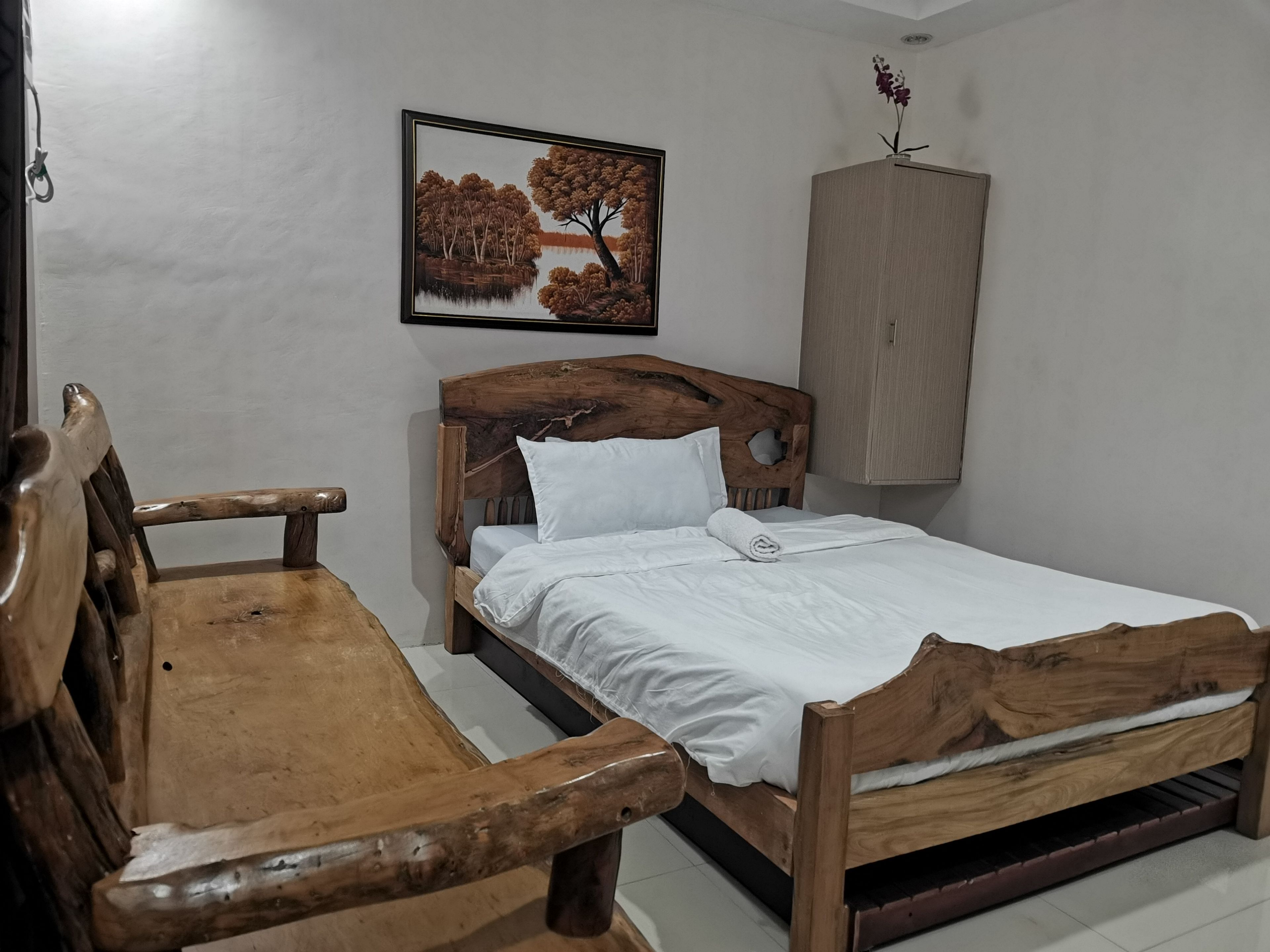 Una cama de madera con ropa de cama blanca, un banco de madera, un armario fijado a la pared y un cuadro con un paisaje de árboles.
