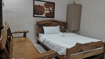 Una cama de madera con ropa de cama blanca, un banco de madera, un armario fijado a la pared y un cuadro con un paisaje de árboles.