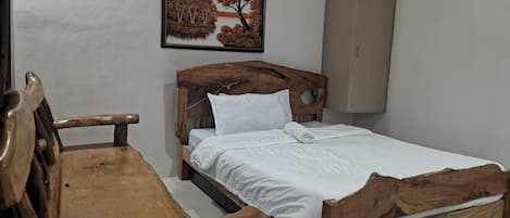 Una cama de madera con ropa de cama blanca, un banco de madera, un armario empotrado en la pared y un cuadro con un paisaje de árboles.