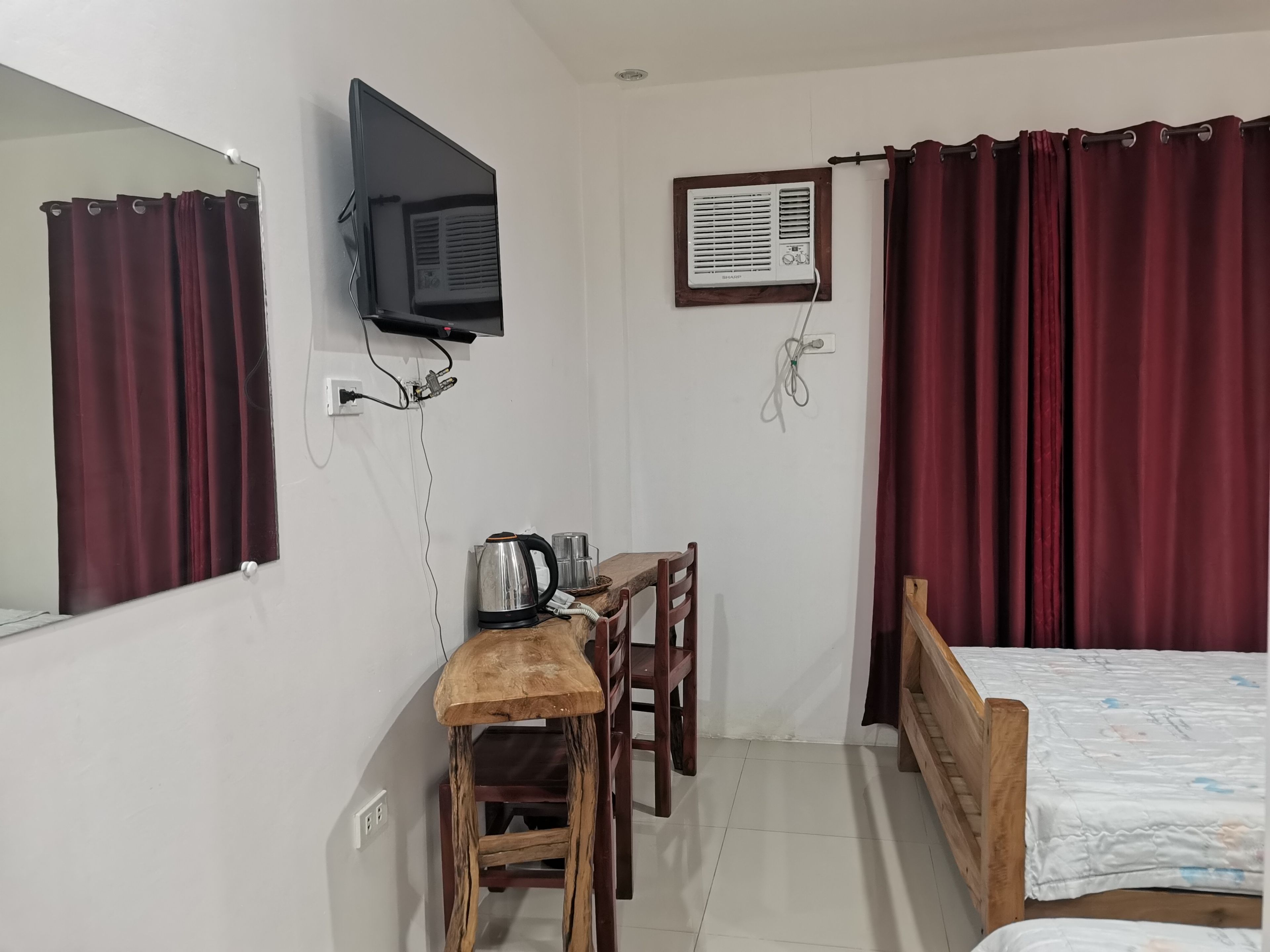 A bedroom with a bed, a desk with a kettle, a chair, a TV mounted on the wall, and an air conditioning unit.