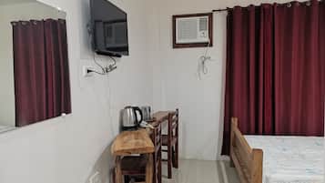 A bedroom with a bed, a desk with a kettle, a chair, a TV mounted on the wall, and an air conditioning unit.
