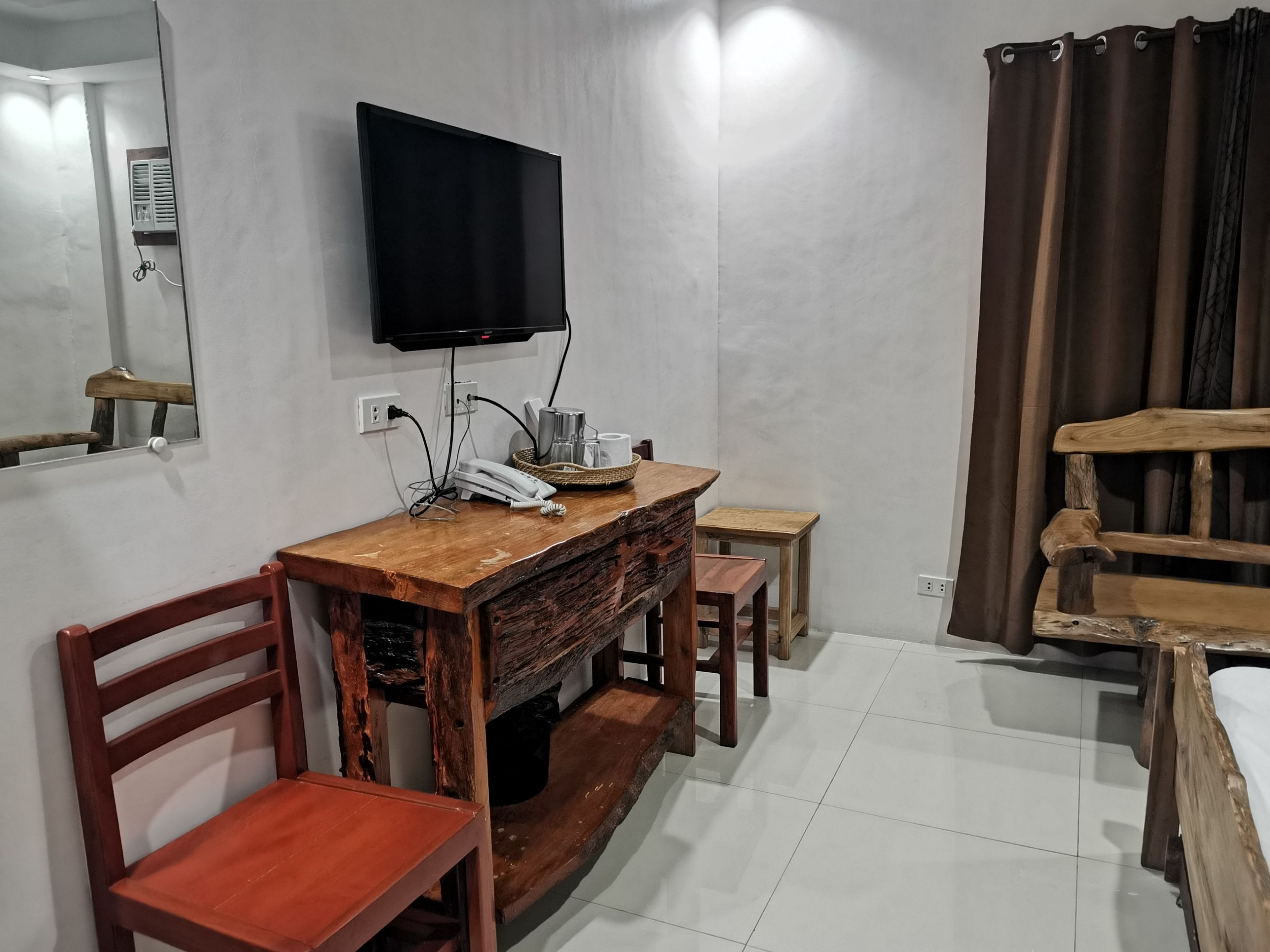 Una habitación con un escritorio de madera, una silla, un televisor montado en la pared, una mesita pequeña y una cama con cabecero de madera.