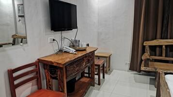 Una habitación con un escritorio de madera, una silla, un televisor montado en la pared, una mesita pequeña y una cama con cabecero de madera.