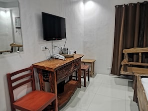 Una habitación con escritorio de madera, televisión montada en la pared, una silla, una mesita y una cama con cabecero de madera.