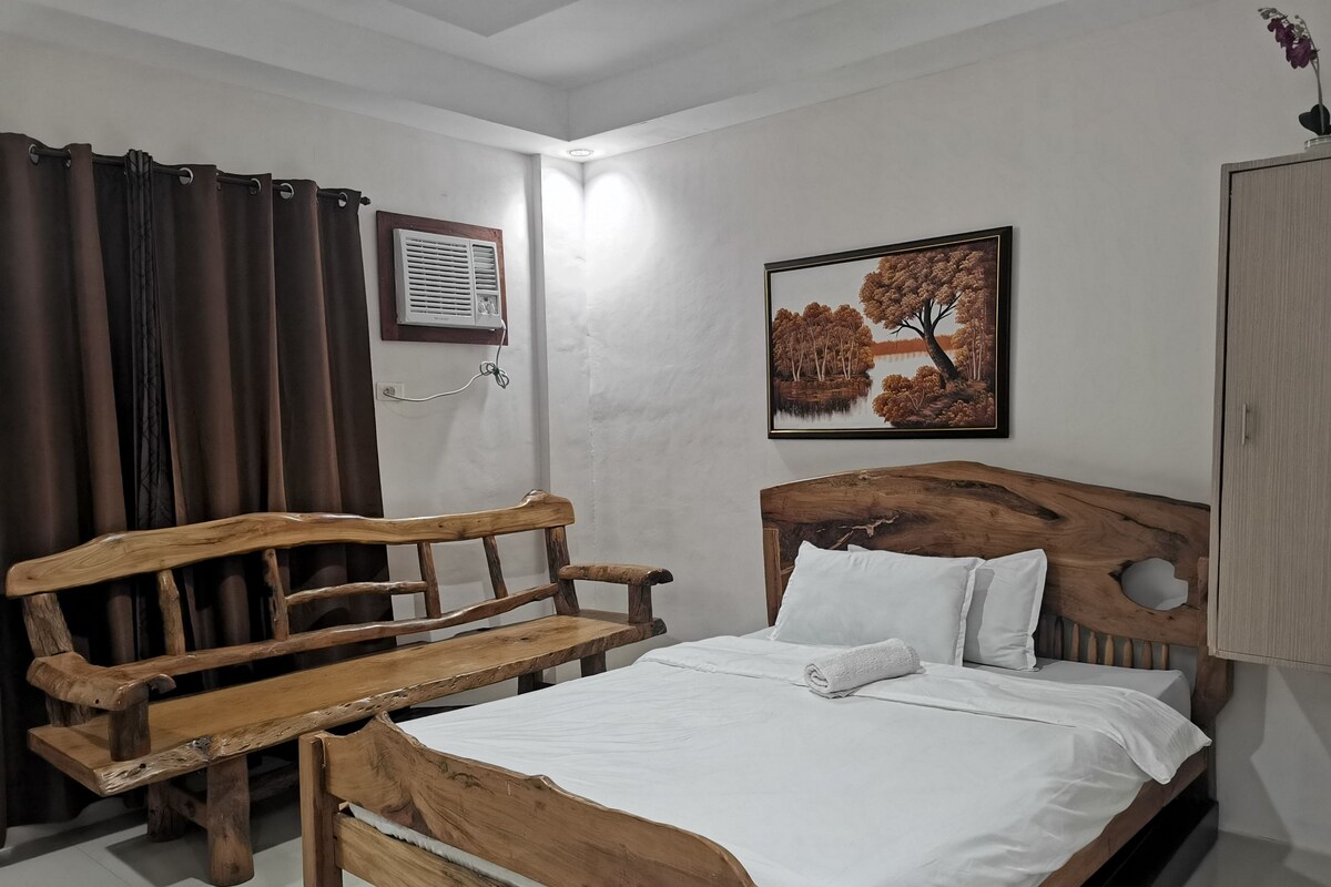 Un dormitorio con una cama y un banco de madera, aire acondicionado de pared y un cuadro con un paisaje de árboles.