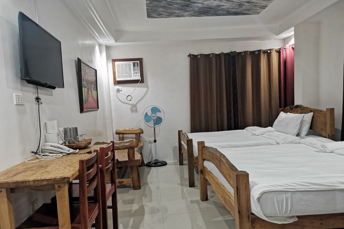 Habitación de hotel con escritorio de madera, dos camas, televisor, ventilador y ventana con cortinas.