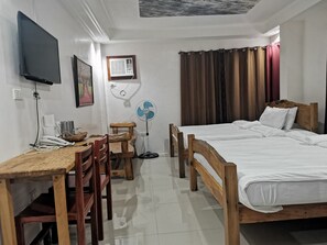 Habitación de hotel con escritorio de madera, dos camas, televisor, ventilador y ventana con cortinas.