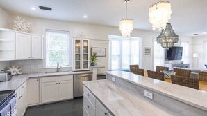 Una cocina moderna con armarios blancos, una isla de mármol y una zona de comedor con mobiliario de mimbre.