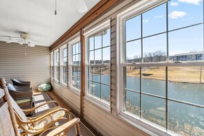 A sunlit porch with wicker chairs, large windows overlooking a body of water and distant buildings.