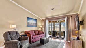 Ein Wohnzimmer mit Sofa, Sessel, Hocker und einem großen Fenster mit Vorhängen.