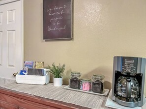 A coffee station with a coffee maker, jars of sugar and creamer, a plant, and a sign on the wall.