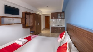 Una camera d'albergo con un letto, una TV a parete, un armadio in legno, uno sgabello e una piccola cucina in fondo.