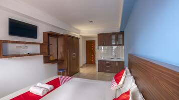 Hotelkamer met een bed, een aan de muur bevestigde televisie, een houten kledingkast, een klein krukje en een kitchenette op de achtergrond.