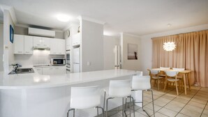 A modern kitchen with white cabinetry, a central island with bar stools, and a dining area with a wooden table and chairs. - Cote D'Azur 4 Sunshine Beach (Sunshine Beach)