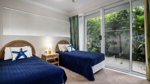 A bedroom with two beds, a nightstand, a lamp, and a sliding glass door leading to a garden. - Cote D'Azur 4 Sunshine Beach (Sunshine Beach)